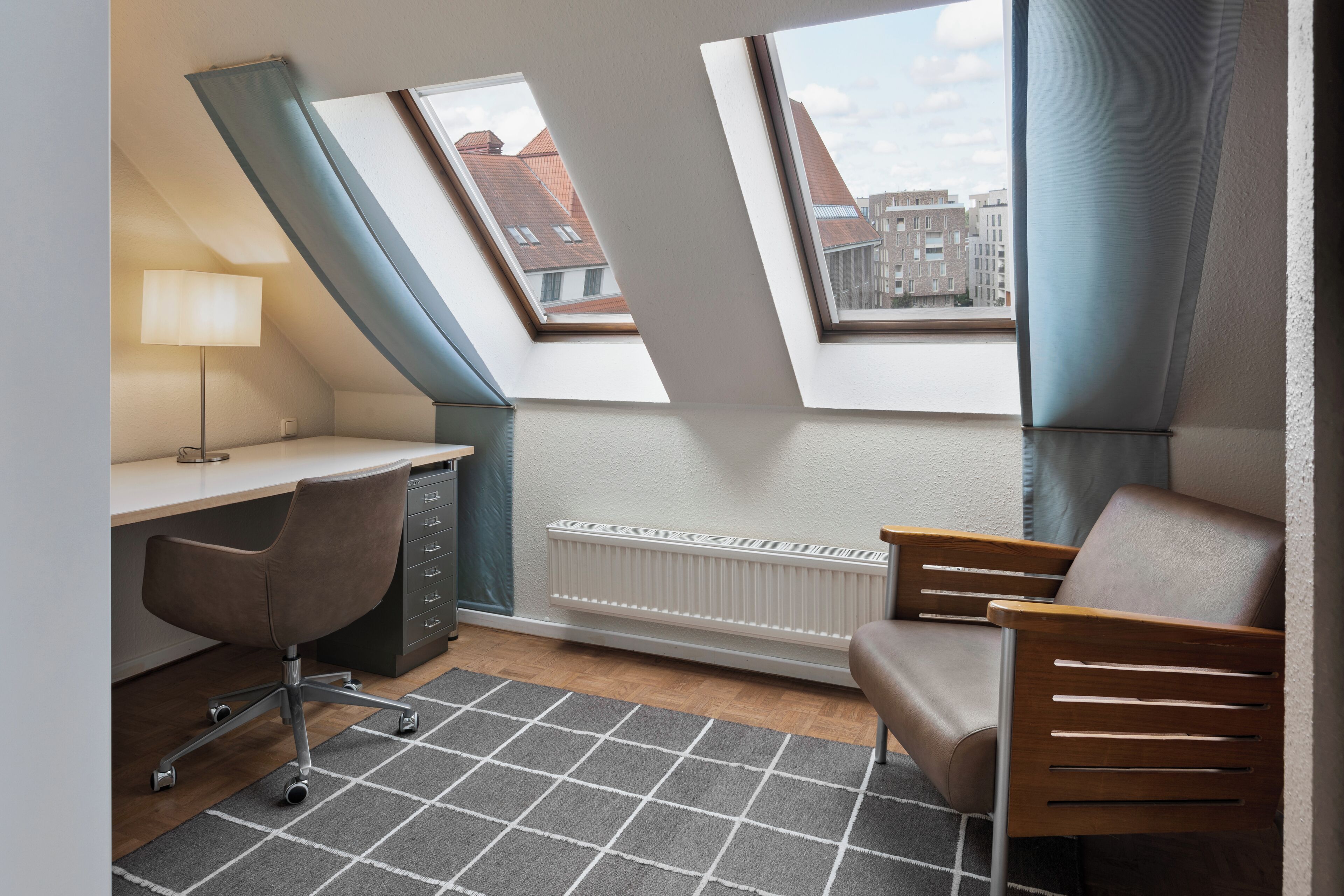 Ein kompakter Raum mit Schreibtisch, Stuhl und Heizkörper. Zwei Dachfenster sorgen für natürliches Licht.