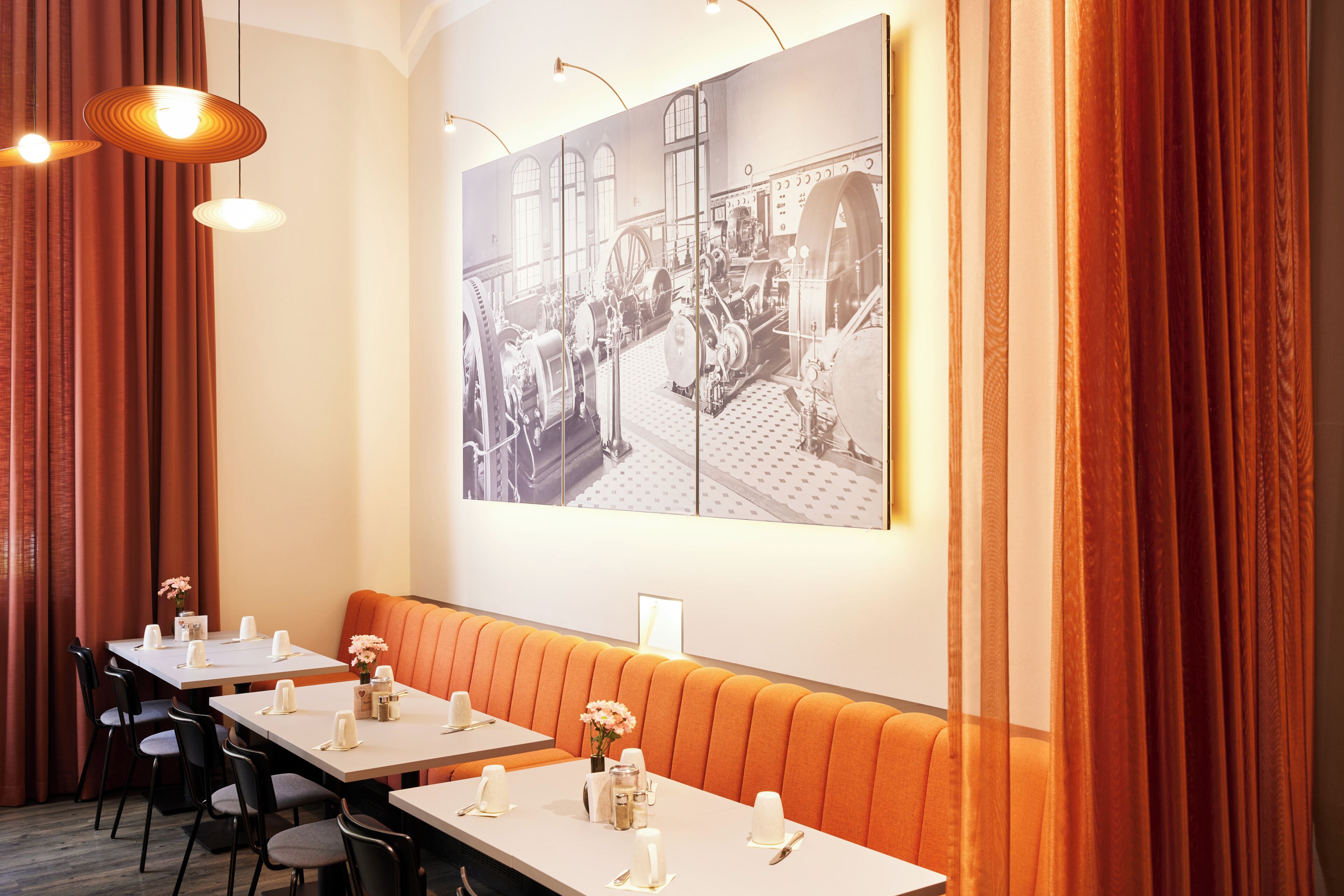 A dining area with tables set for service, a long orange booth, and a large black-and-white industrial photo on the wall.