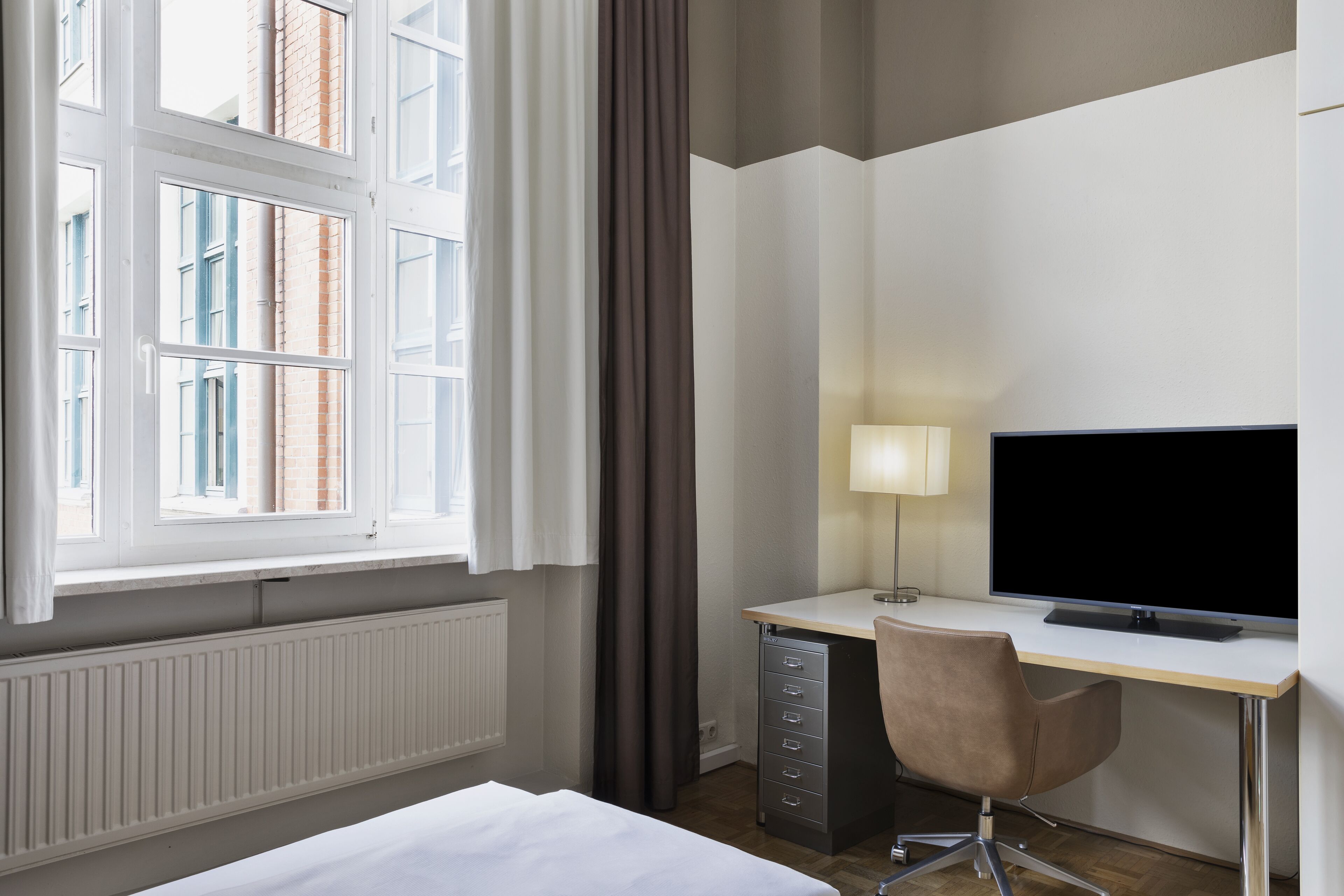 Ein Hotelzimmer mit einem Bett, einem Schreibtisch mit Computer und Lampe, einem Sessel und einem Fenster mit Vorhängen.