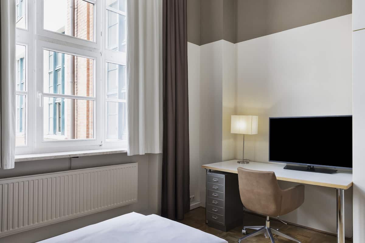 Ein Hotelzimmer mit einem Bett, einem Schreibtisch mit Computer und Lampe, einem Sessel und einem Fenster mit Vorhängen.