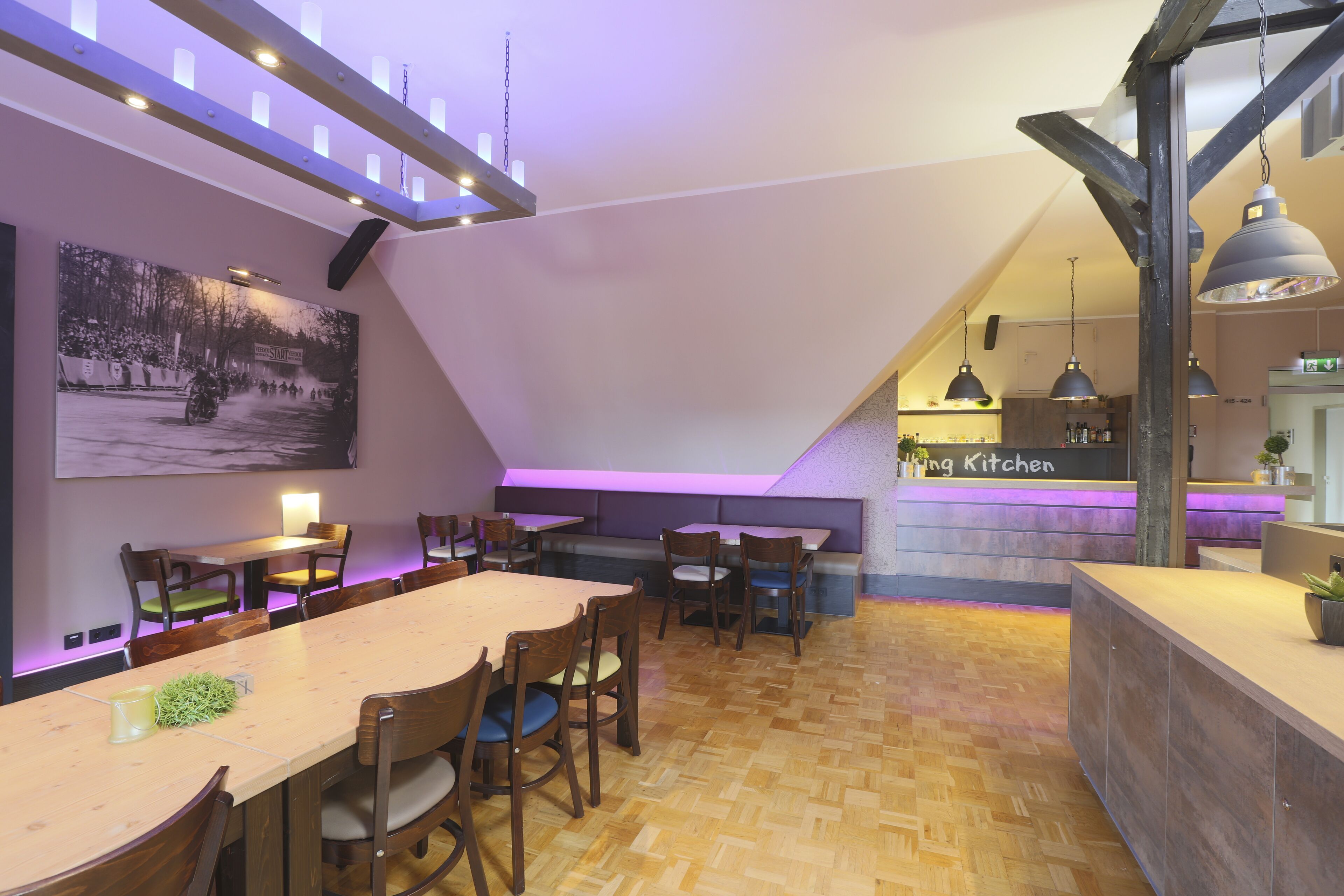 A dining area with wooden tables and chairs, a kitchen in the background, and a large painting on the wall.