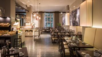 A spacious restaurant with multiple dining tables and chairs, a counter with baked goods, and a large window providing natural light.