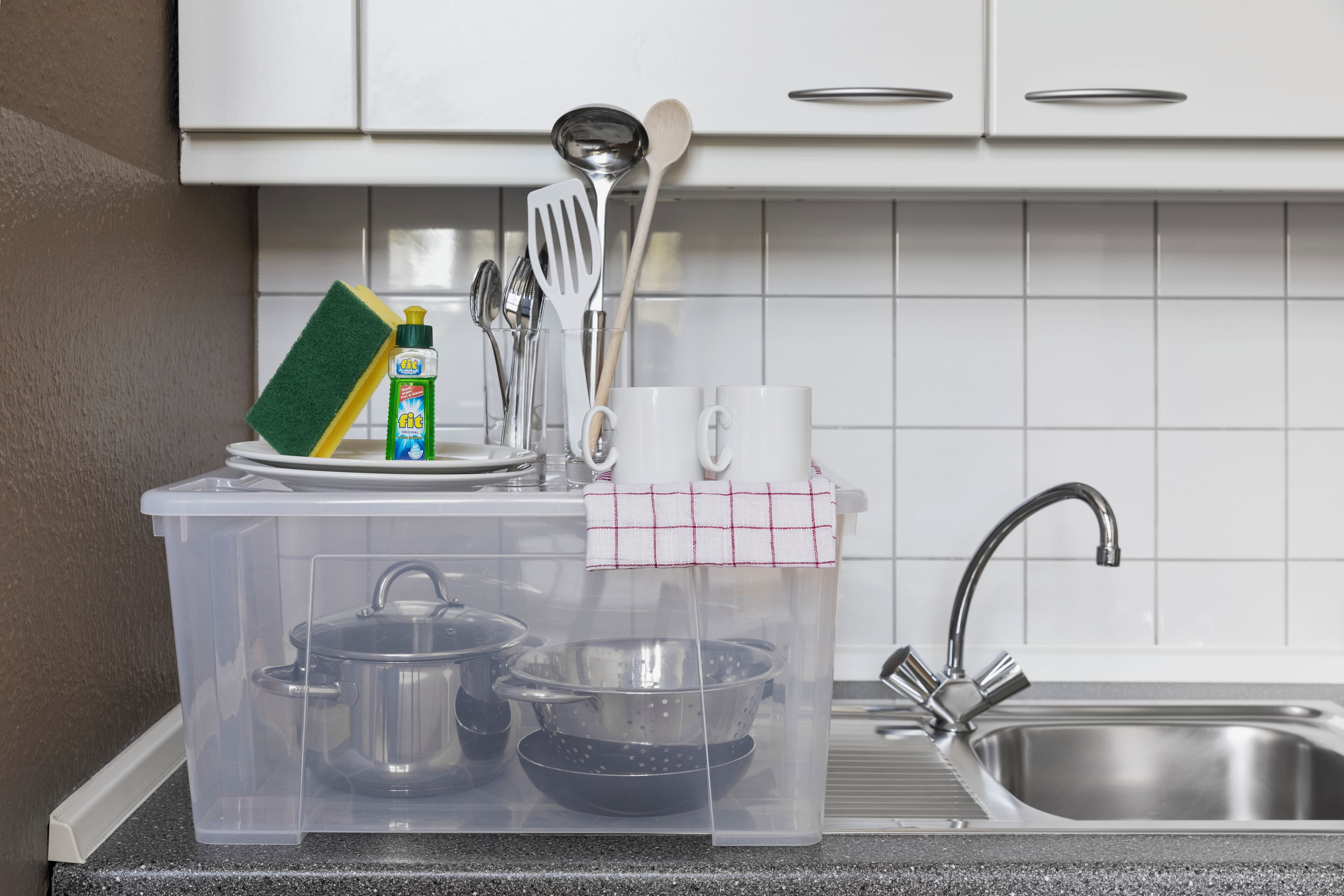 A kitchen with white cabinets, a stainless steel sink, and a plastic storage organizer containing kitchen utensils and dishes.