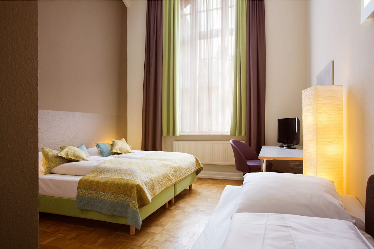 Ein Hotelzimmer mit zwei Betten, einem Schreibtisch mit Computer und einem Fenster mit Vorhängen.