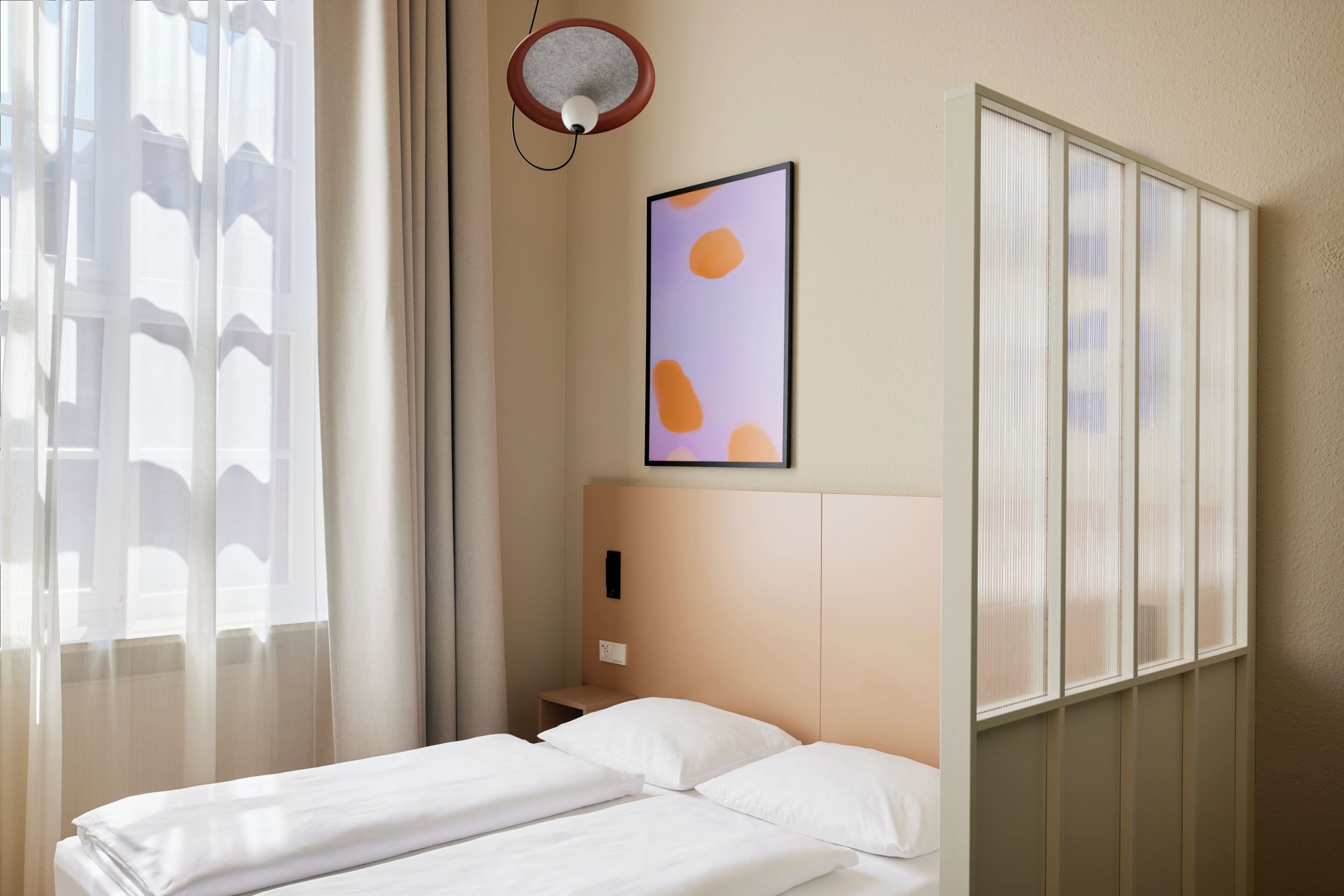 A hotel room with two beds, a bedside table, a wall-mounted light, and a window with sheer curtains.