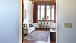 A room with a wooden ceiling, a window with red curtains, a white sofa, a patterned rug, and a wooden chair. - Maggio View Apartment (Firenze)