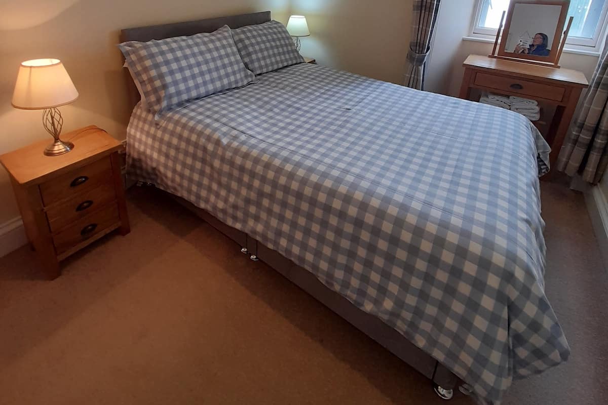 Une chambre à coucher avec un lit, une table de chevet, une lampe, une fenêtre avec des rideaux et un miroir sur une commode.
