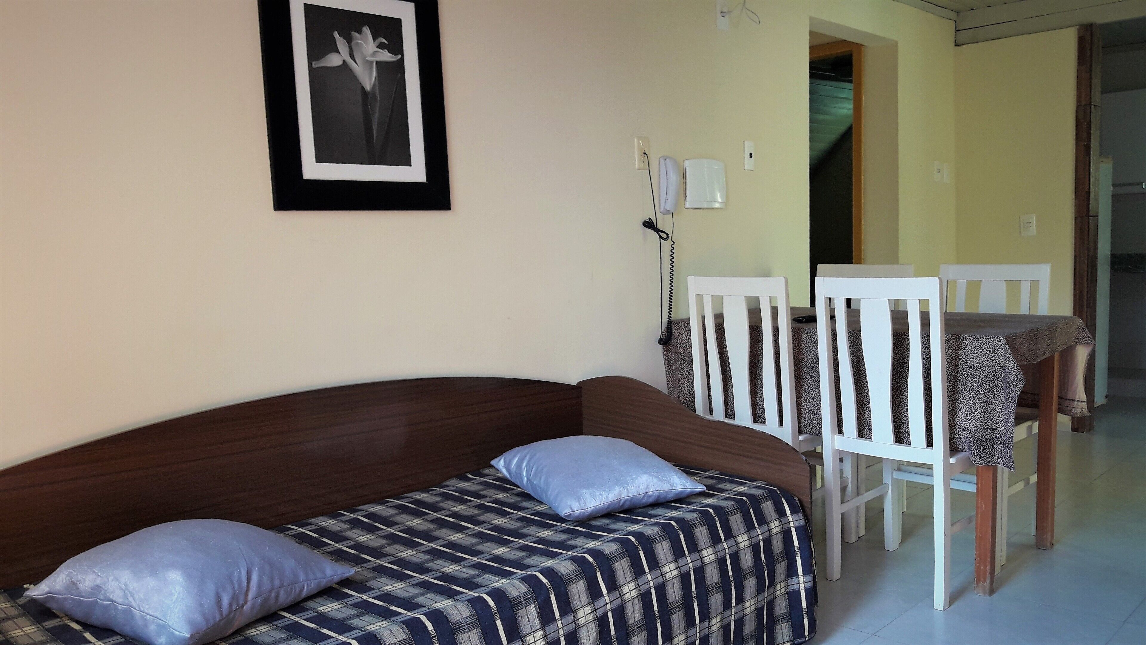 Una cama individual con colcha escocesa y dos almohadas, una silla blanca, una mesita con mantel estampado y un cuadro en la pared.