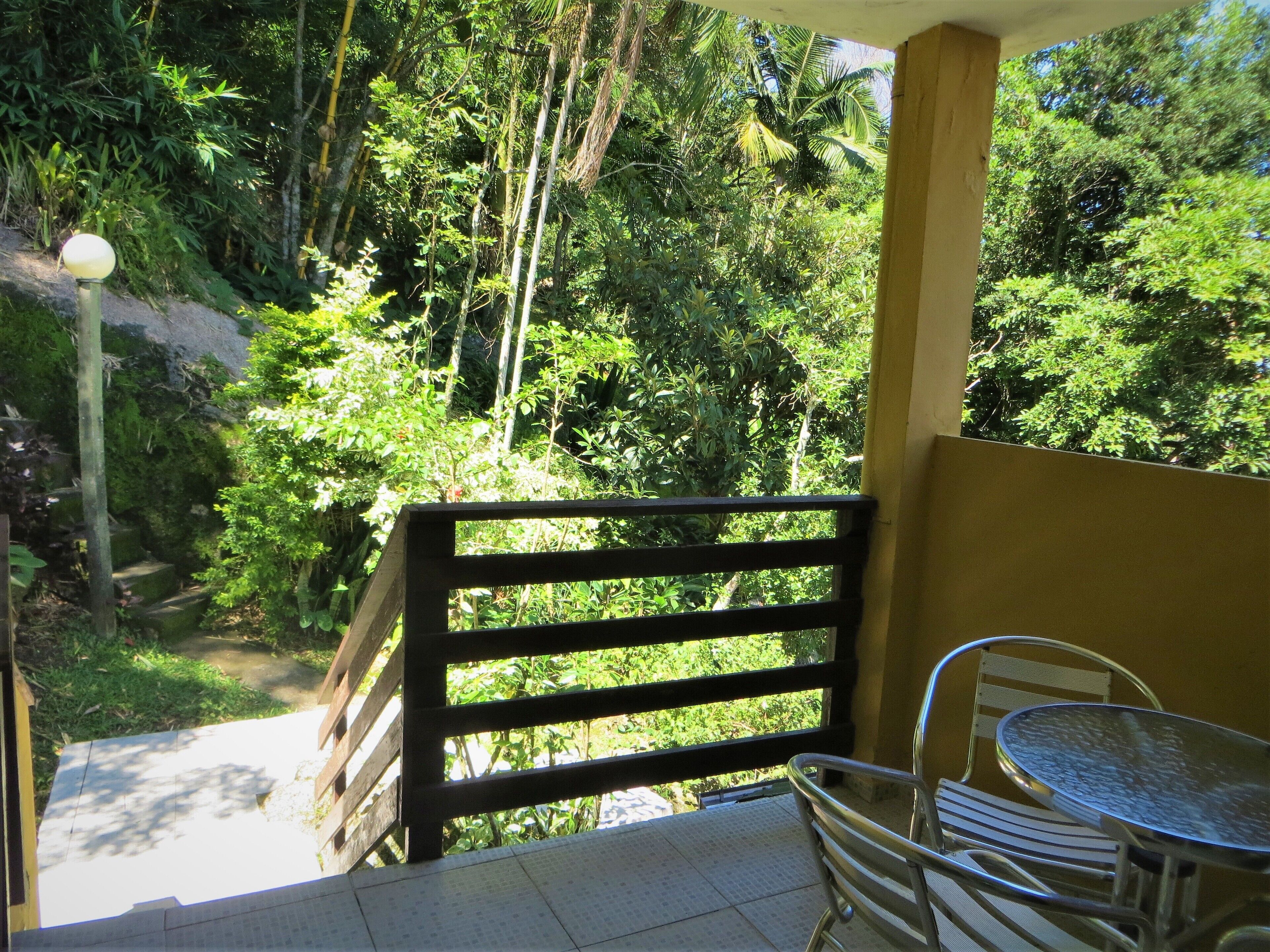 A balcony with a metal table and chairs, overlooking a lush garden with various plants and trees.