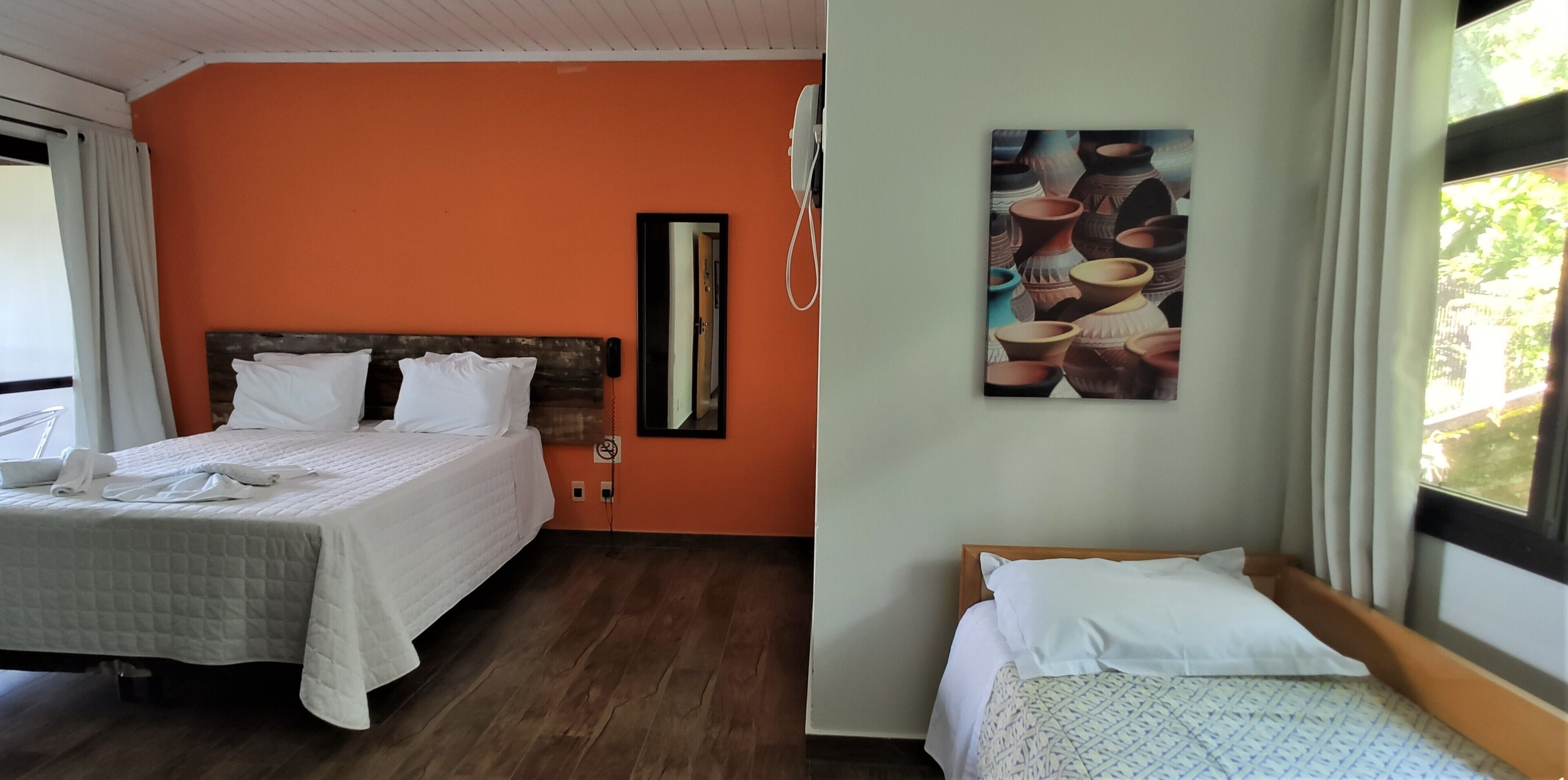 A hotel room with two beds, an orange wall, a wooden headboard, a painting of pottery, and a window with curtains.