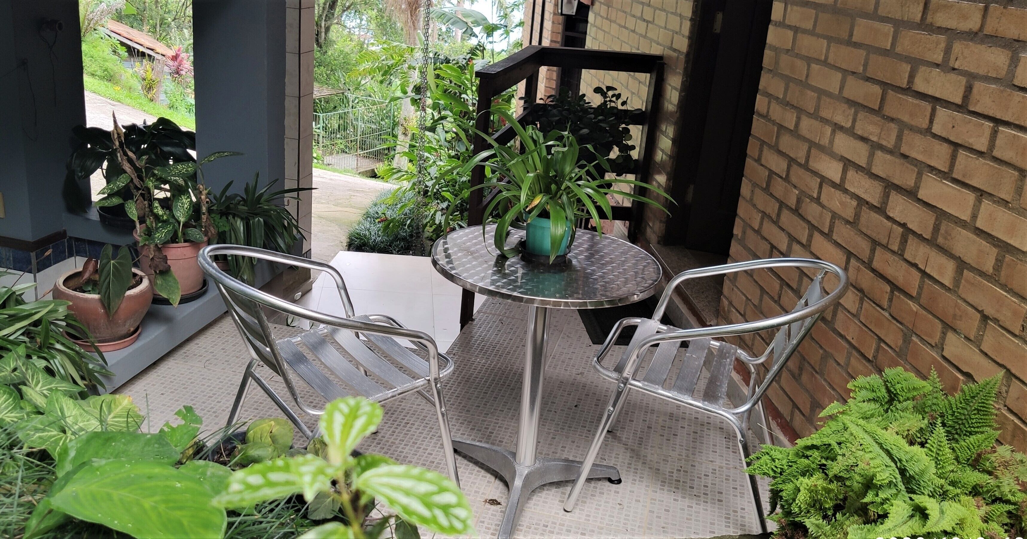A small patio area with a round metal table and two metal chairs, surrounded by potted plants and a brick wall.