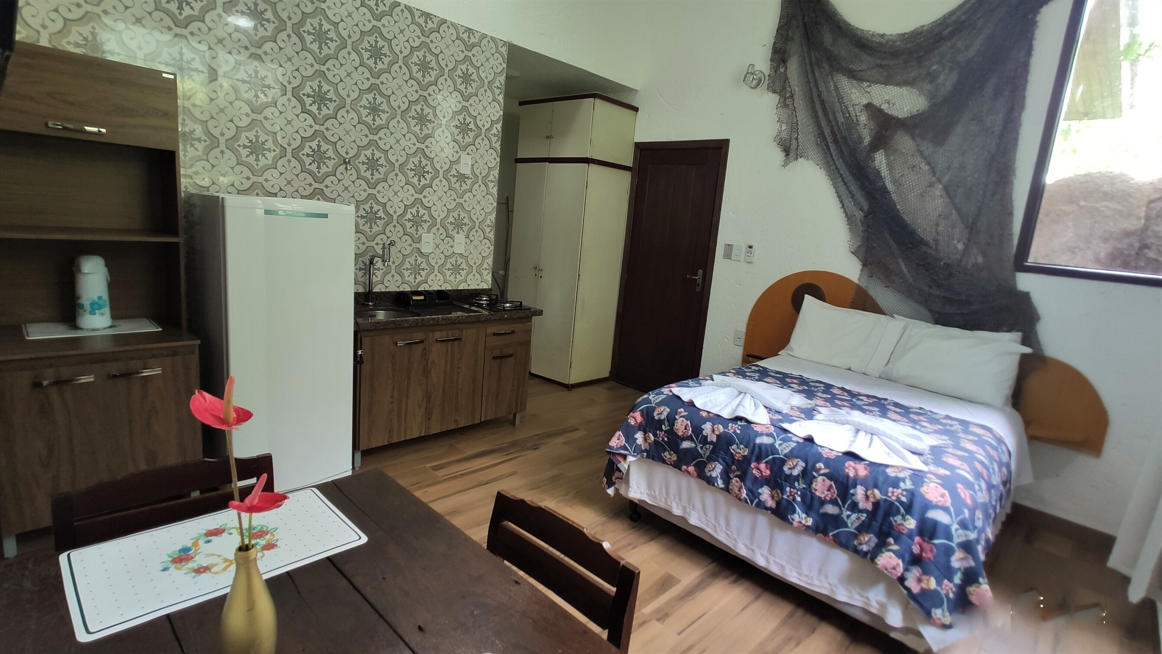 A bedroom with a bed, a desk, a chair, a small table, and a refrigerator.
