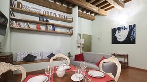 Een eetgedeelte met een houten tafel gedekt voor vier personen, een boekenkast met boeken en decoratieve items, een grijze bank en een lijst met kunstwerk aan de muur.