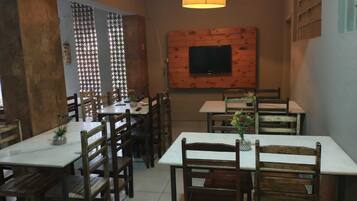 Um restaurante com mesas e cadeiras de madeira, uma TV presa a uma parede de madeira e um lustre pendurado.