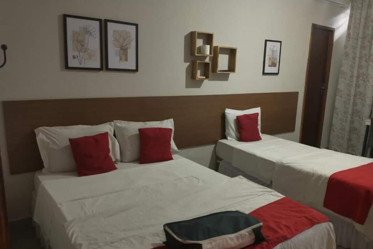 Habitación de hotel con dos camas, cada una con ropa blanca y almohadas rojas. En la pared hay cuadros enmarcados y una estantería de madera con una taza y una caja de pañuelos.