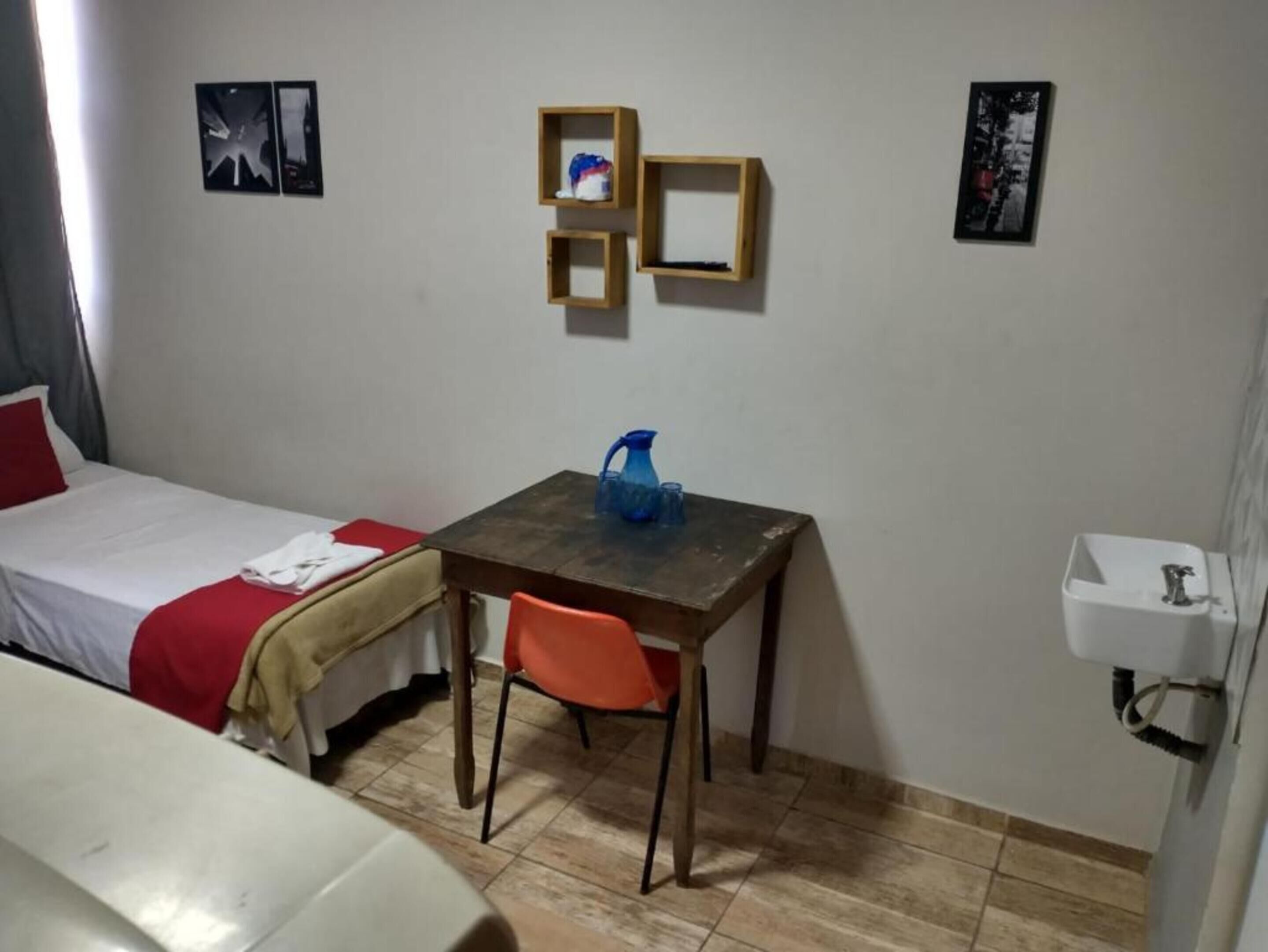 A small room with a bed, a table, and a chair. There are pictures on the wall and a sink in the corner.