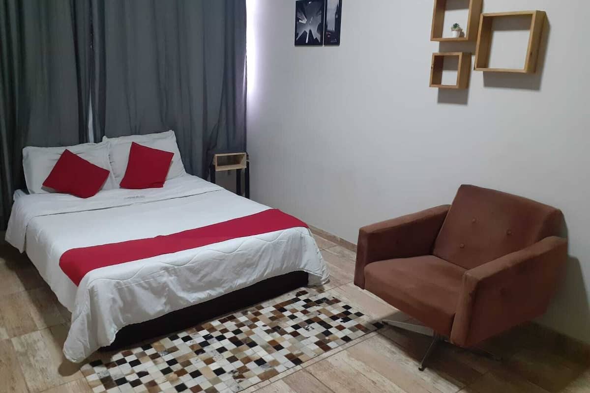 Un dormitorio con cama, una silla, una alfombra a cuadros y una pared con cuadros enmarcados.