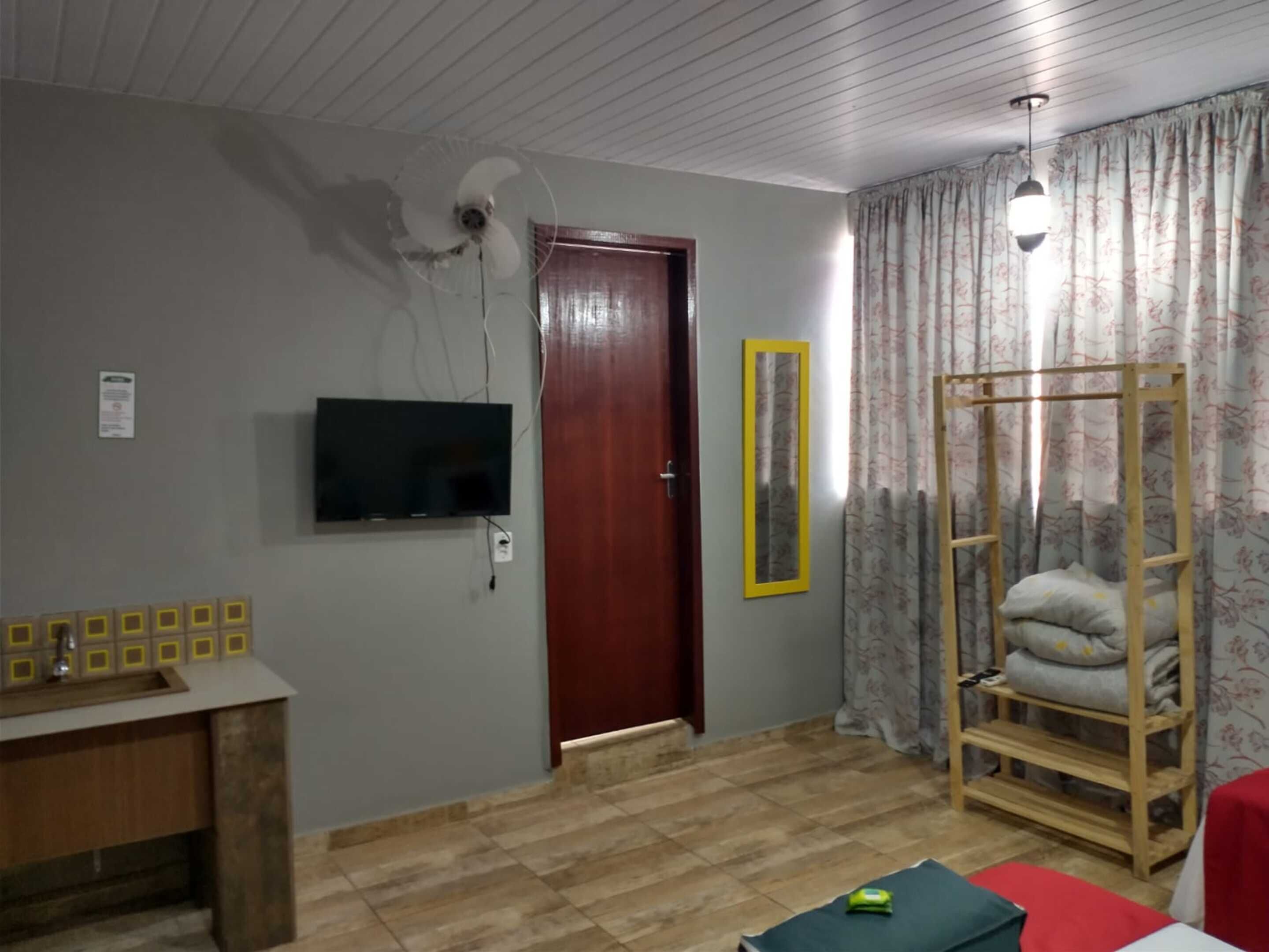 A room with a wooden desk, a television mounted on the wall, a door, a mirror, a wooden shelf with pillows, and a ceiling fan.