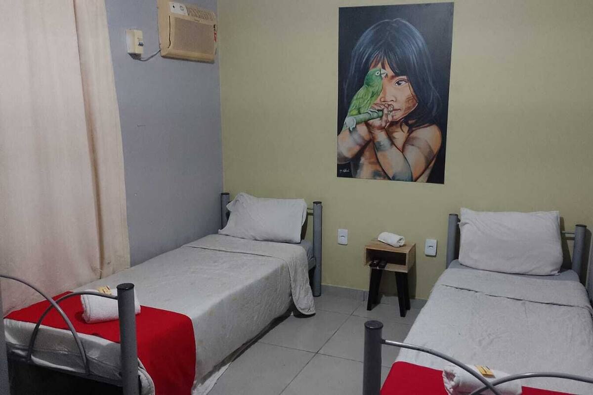 Habitación con dos camas, cada una con colcha roja y blanca, una mesita de noche con lámpara y un aire acondicionado instalado en la pared.