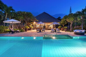 A swimming pool with lounge chairs and umbrellas, surrounded by tropical plants and a building with a tiled roof. - Private Beach front Villa 12p: Accomplish your body and soul! (Dencarik)