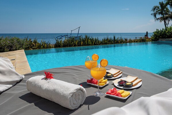 A poolside setting with a towel, fruit, and drinks on a table, overlooking the sea.