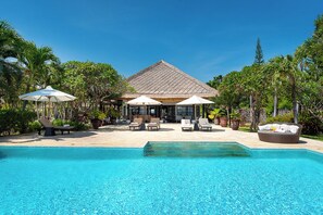 A pool area with lounge chairs and umbrellas, surrounded by lush greenery and a building with a tiled roof.