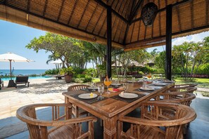An outdoor dining area with a thatched roof, wooden tables, and chairs, overlooking a pool and lush greenery.