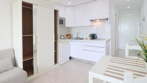 Cozinha moderna com armários brancos, micro-ondas embutido e um frigorífico pequeno. Há uma mesa de jantar com cadeiras e uma planta à direita.