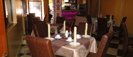 Un espace repas avec des tables disposées pour quatre personnes, chacune agrémentée d’une bougie, d’assiettes et de couverts. Les chaises sont recouvertes d’un cuir marron foncé. Le sol présente un motif à carreaux, et les murs sont peints dans une teinte chaude.