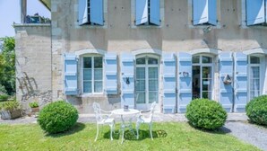 A stone building with light blue shutters, a small garden with a table and chairs, and a gravel path.
