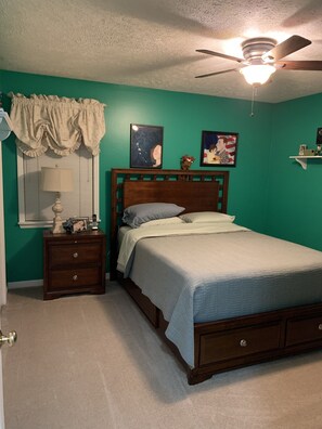 A bedroom with a bed, a nightstand, a lamp, a ceiling fan, and two framed pictures on the wall.