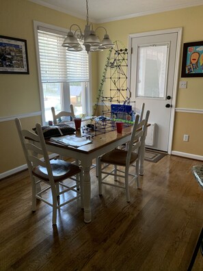 A dining area with a wooden table and white chairs, a chandelier, and a window with blinds.