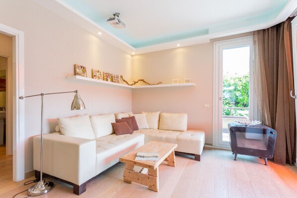 A modern living room with a sectional sofa, a coffee table, a floor lamp, and a TV stand. There are shelves with decorative items above the sofa.