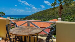 A balcony with a round table and chairs, overlooking a coastal landscape with a view of the sea and a hill with vegetation.