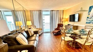 Une chambre d’hôtel avec un canapé en cuir, une table à manger avec des chaises, un téléviseur à écran plat et une grande fenêtre avec des rideaux.