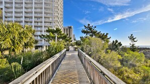 Une promenade en bois mène à un immeuble de grande hauteur doté de balcons, entouré de verdure et surplombé d’un ciel dégagé.