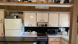 Een kleine keuken met witte kastjes, een koelkast, een fornuis, een magnetron en een prullenbak.