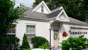 Une maison blanche avec un porche à l’avant, un petit jardin avec des fleurs et un abreuvoir pour les oiseaux, et une guirlande sur la porte.