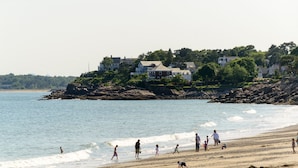 Une plage où l’on voit des gens se promener et jouer, des maisons perchées sur une falaise rocheuse et un ciel dégagé.