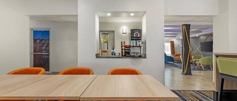 A conference room with a wooden table and orange chairs, a coffee station with two coffee machines, and a view of a cityscape through a glass door.