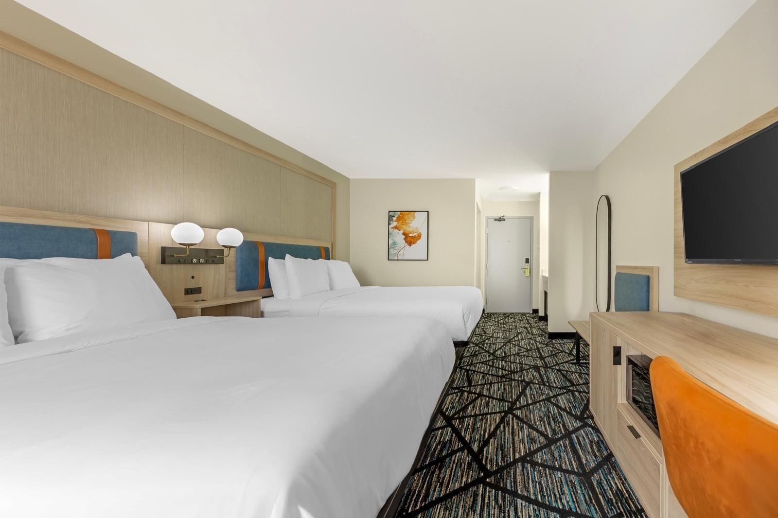 A hotel room with two beds, a desk, a flat-screen TV, and a patterned carpet.