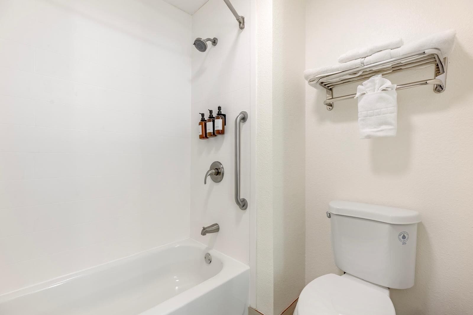 Un baño con una bañera blanca, un inodoro y una ducha con cabezal de ducha manual. Hay artículos de aseo en la pared y un toallero con una toalla.