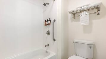 Un baño con una bañera blanca, un inodoro y una ducha con cabezal de ducha manual. Hay artículos de aseo en la pared y un toallero con una toalla.