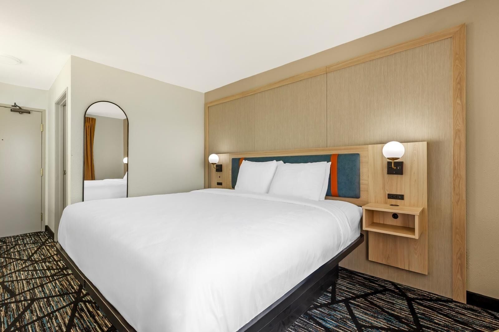 A hotel room with a large bed, a bedside table with a lamp, a mirror, and a carpeted floor.
