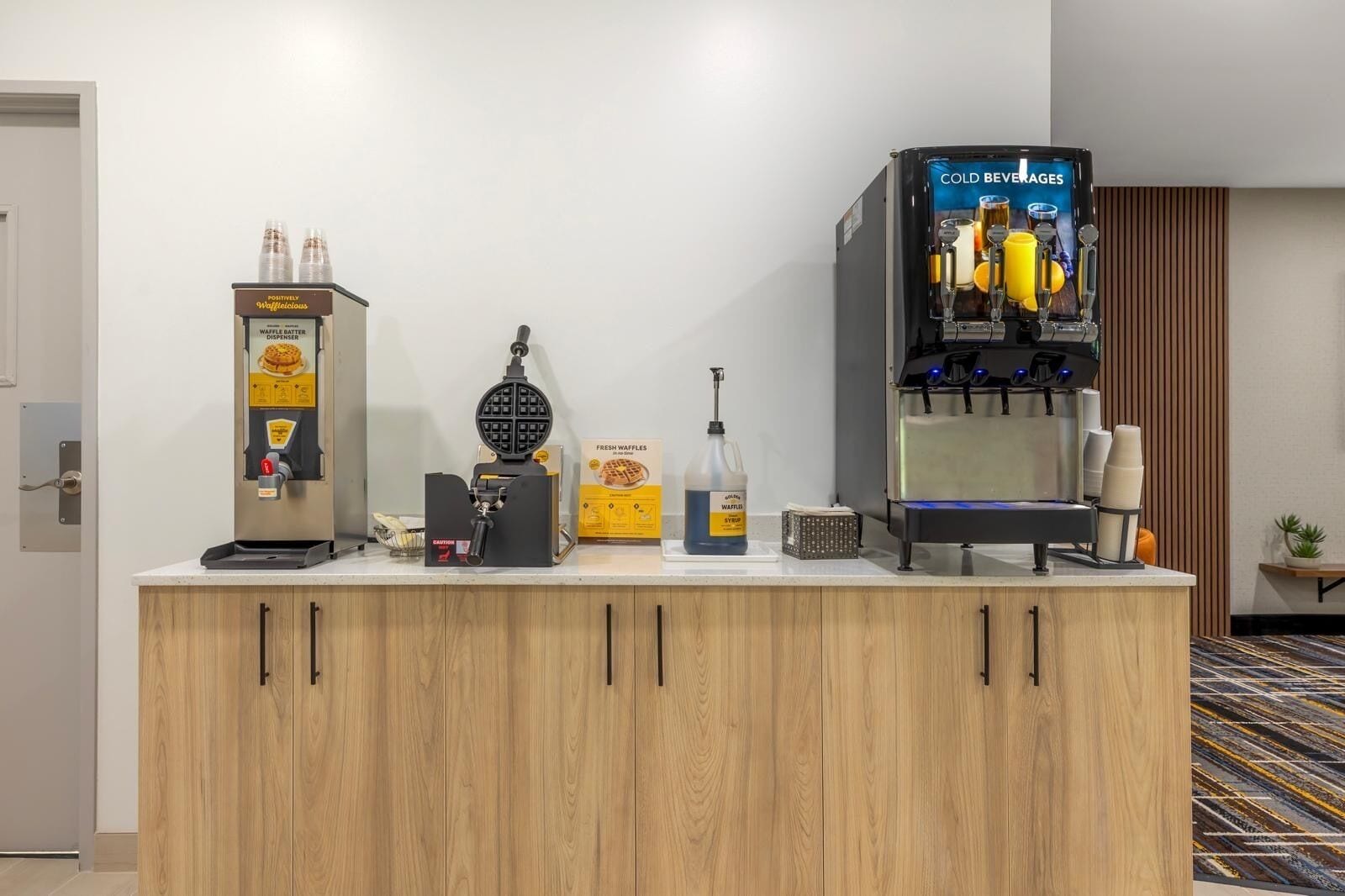 Une petite cuisine équipée d’un appareil à gaufres, d’un distributeur de pâte à gaufres, d’un flacon de sirop pour gaufres et d’un distributeur de boissons fraîches.