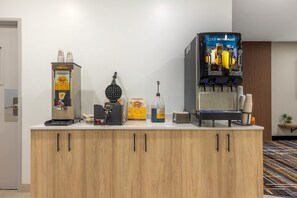 A kitchenette with a waffle maker, a waffle batter dispenser, a waffle syrup bottle, and a cold beverage dispenser. - Bay Inn & Suites Troy (Troy)