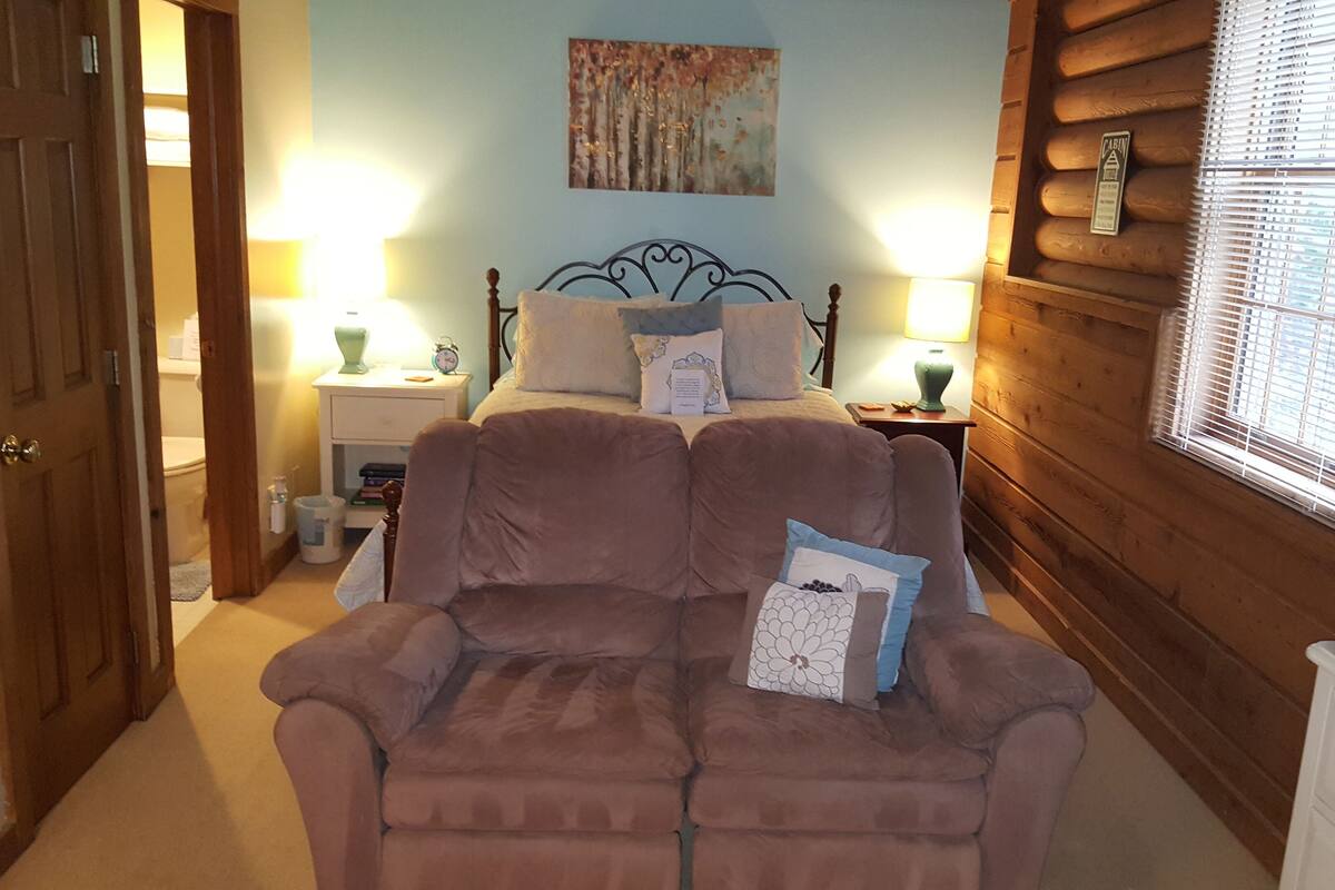 A cozy bedroom with a double bed, a wooden headboard, a nightstand with a lamp, and a window with blinds.