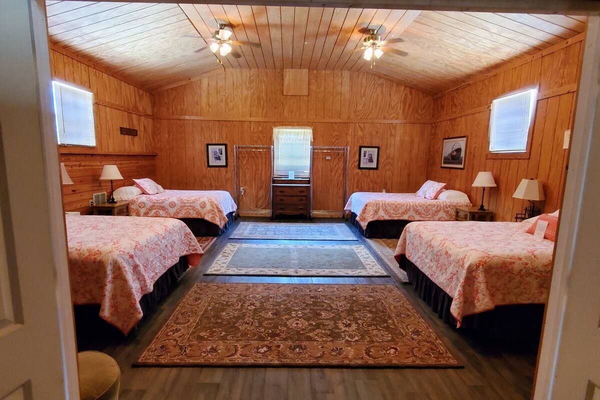A room with wooden walls and ceiling, two beds with floral bedding, a central rug, and a ceiling fan.
