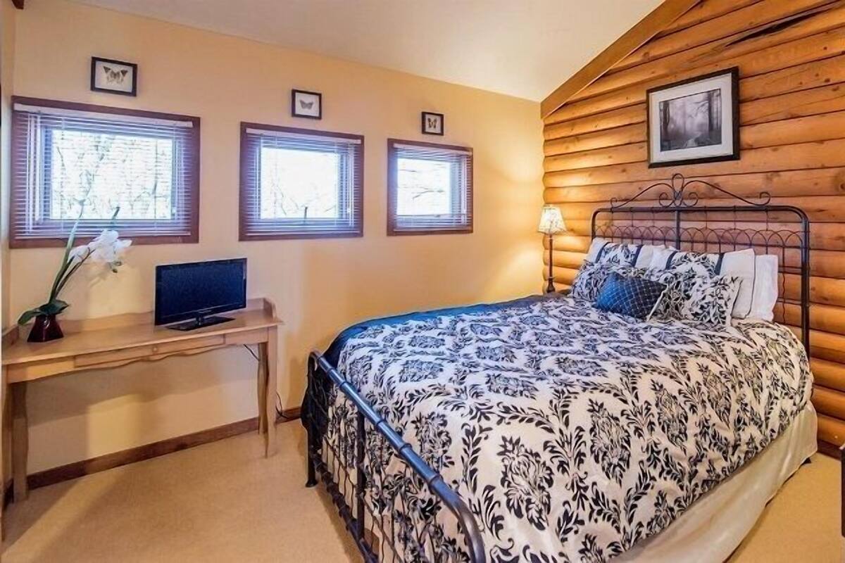 A bedroom with a wooden sloped ceiling, a bed with patterned bedding, a desk with a computer, and a lamp.