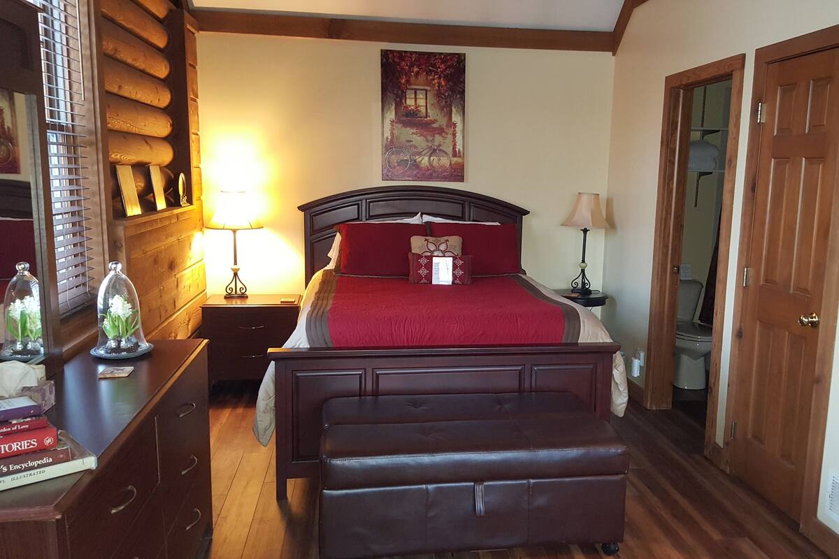 A bedroom with a wooden headboard, a red bedspread, a nightstand with a lamp, a dresser with books, and a wooden bench.